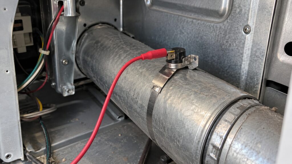 A small temperature sensor clipped to the exhaust duct inside the back of a dryer with a meter lead on its terminal