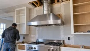 A wide shot of a stainless steel LG range hood mounted above a gas range in a kitchen under renovation, with round metal ductwork visible running up through an open ceiling cavity