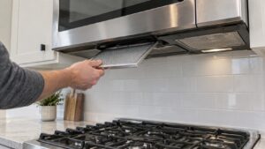 A stainless steel LG over-the-range microwave above a gas cooktop, a hand sliding a metal grease filter out from the underside