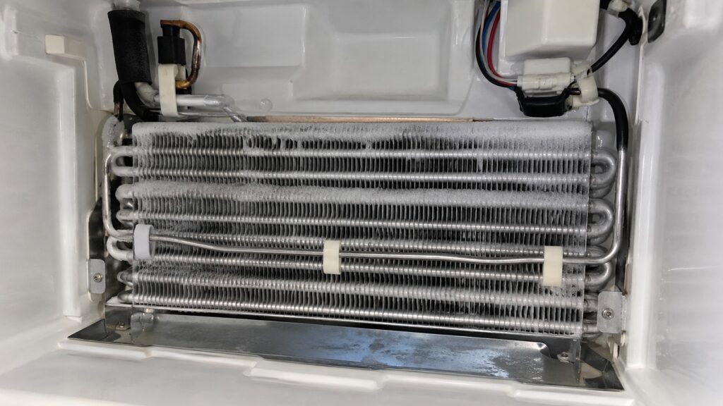 A refrigerator evaporator coil with a thin layer of frost and a defrost heater element running along its fins, seen with the rear panel removed